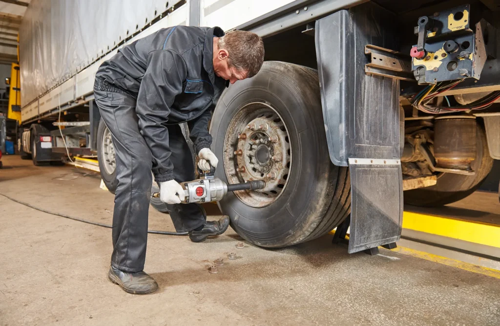 Commercial truck mechanic using a pneumatic impact wrench to tighten or loosen the lug nuts on a large semi-trailer truck wheel in a repair garage.