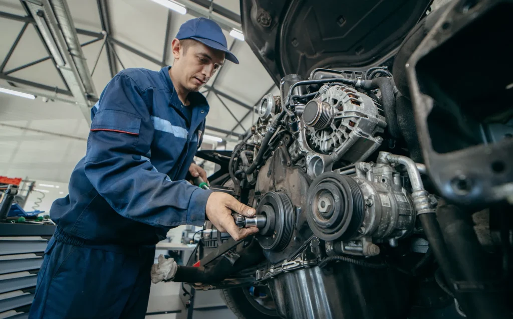 Professional mechanic in a blue uniform inspecting the engine of a car, focusing on the alternator and pulleys in an auto repair shop.