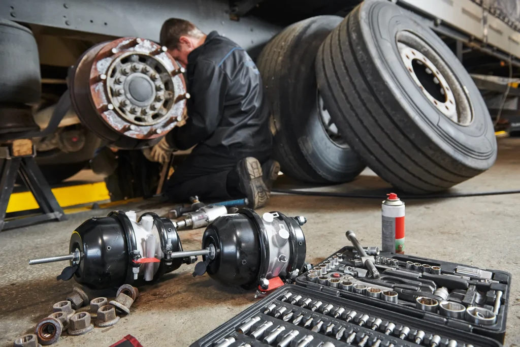 Truck mechanic repairing or replacing the brake drum and axle on a heavy vehicle, with new air brake chambers and tools in the foreground.